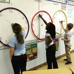 Children in a classroom using large wall clocks to practice telling time.